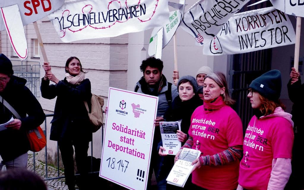 (Photo Rights: Wir packen's an) 7 persons standing next to each other infront of a beige wall with black and pink clothing holding different banners with slogans such as 